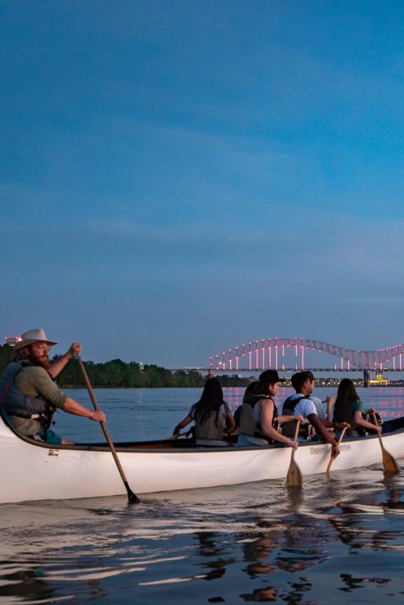 A group of people row a multi-person canoe on the Mississippi River with an illuminated bridge in the background. 