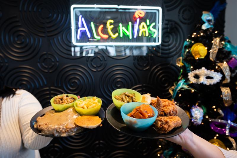 two people holding plate of soul food by Alcenia's neon sign
