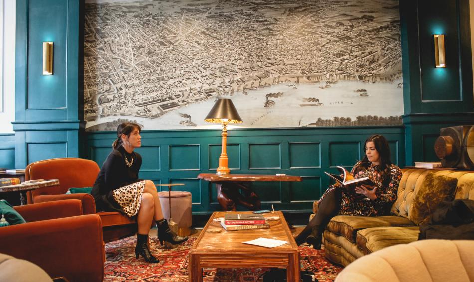 Two women sitting inside the lobby of Arrive Hotel with a large throwback illustration of the Mississippi riverfront on the wall behind them.